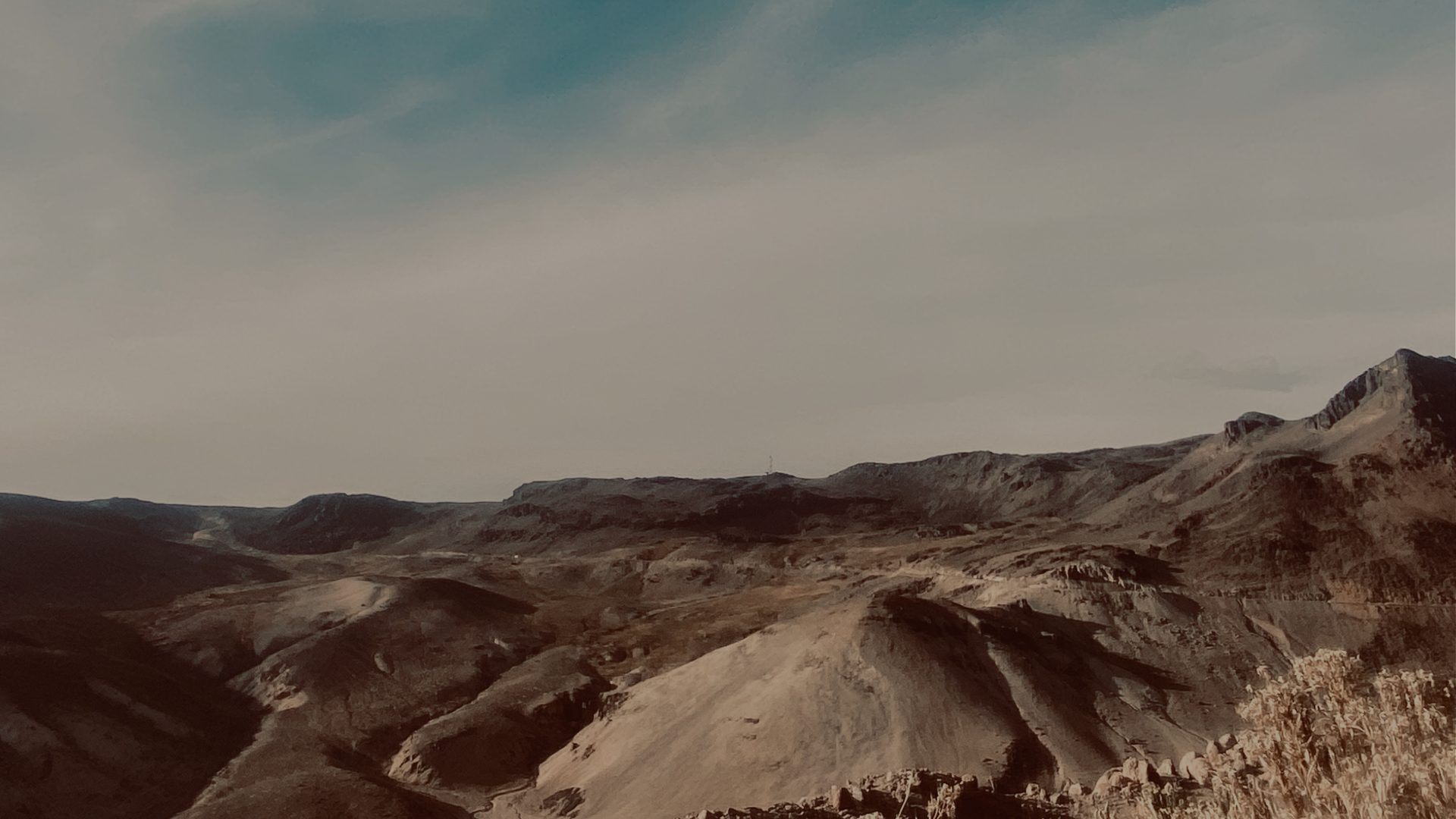 A wide view over arid, layered mountain ridges in muted browns, receding toward a distant range under a pale, washed-out sky.