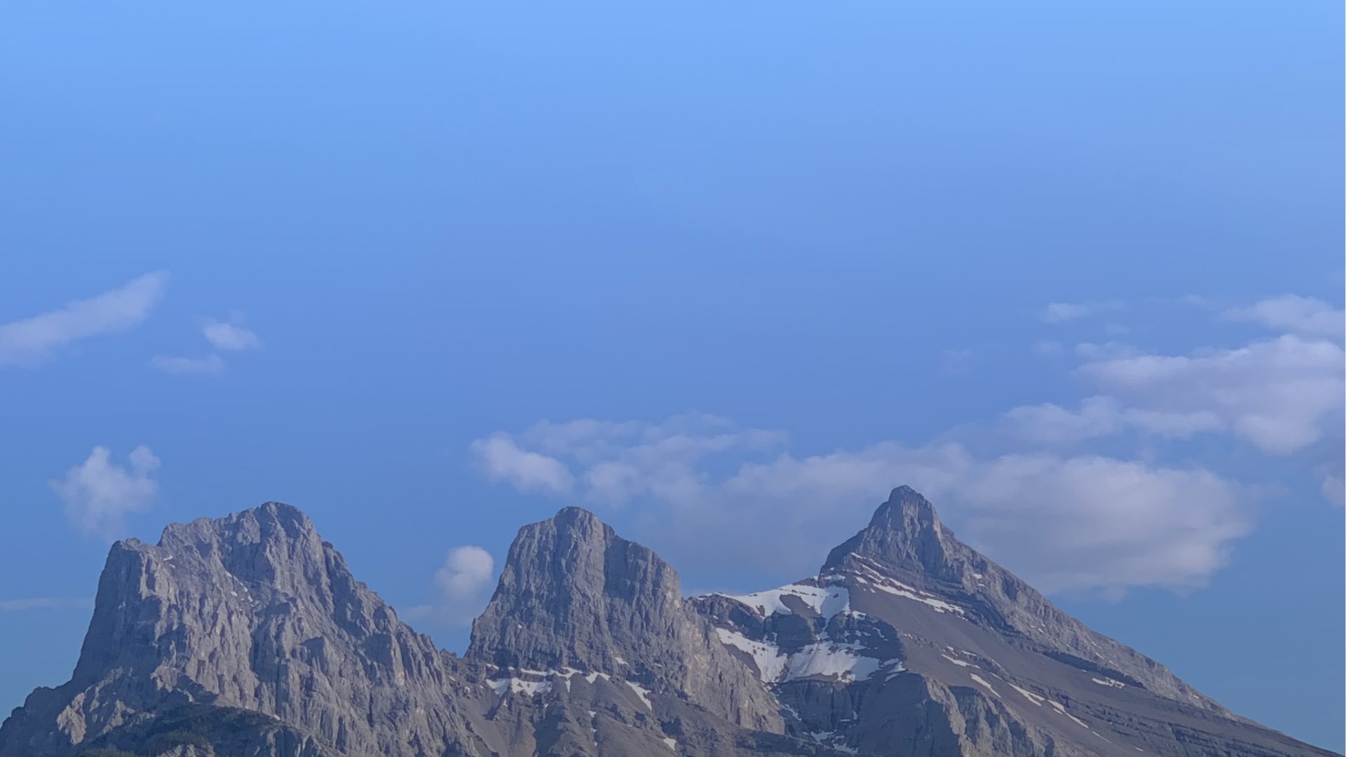 Three jagged, snow-streaked mountain peaks rising against a clear blue sky with scattered clouds along the ridgeline.